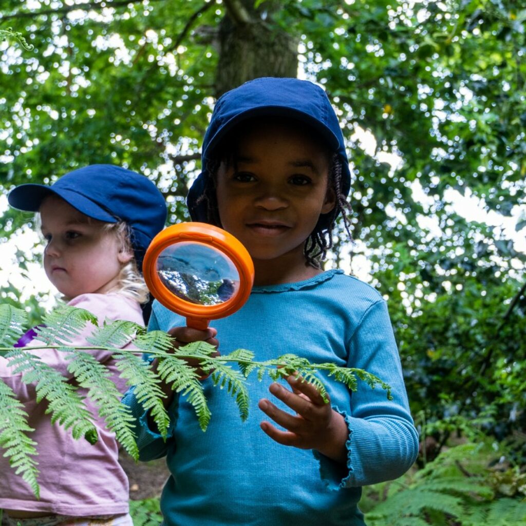 two Kindergarden pupils holding magnifying glasses and looking at plants during a Forest School session at St Mary's Essex