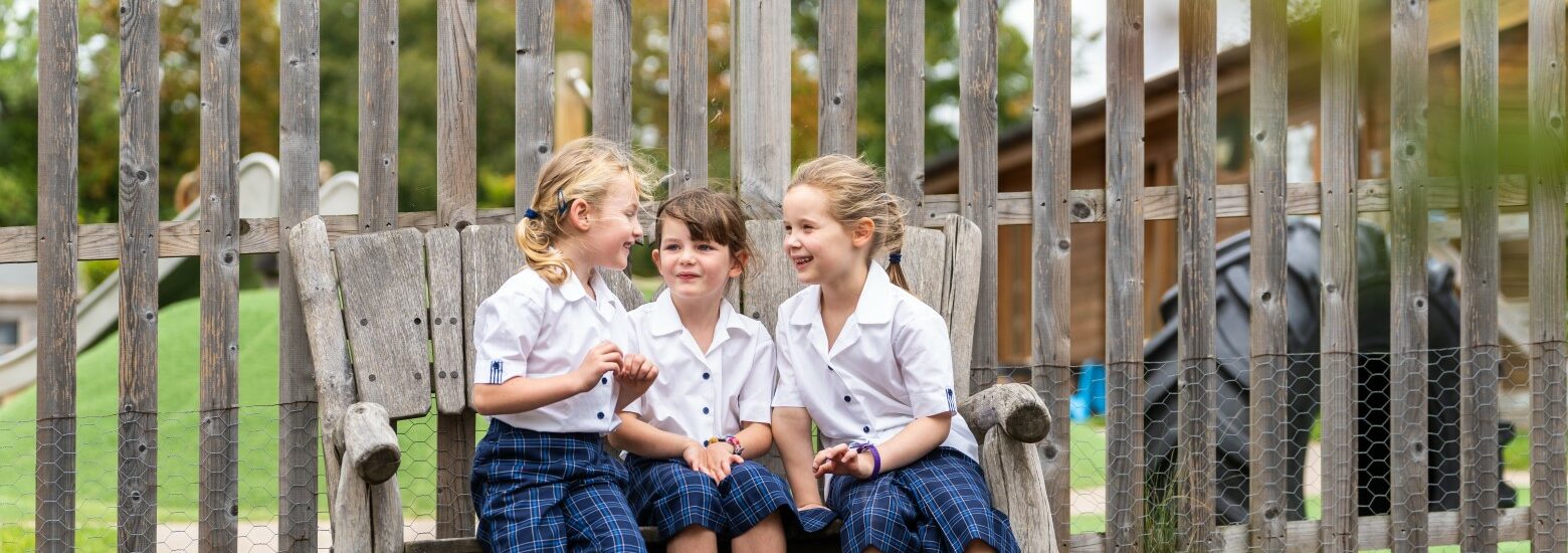 Three lower school girls sitting in an outdoor bench talking excitedly while wearing wellies at St Mary's in Essex