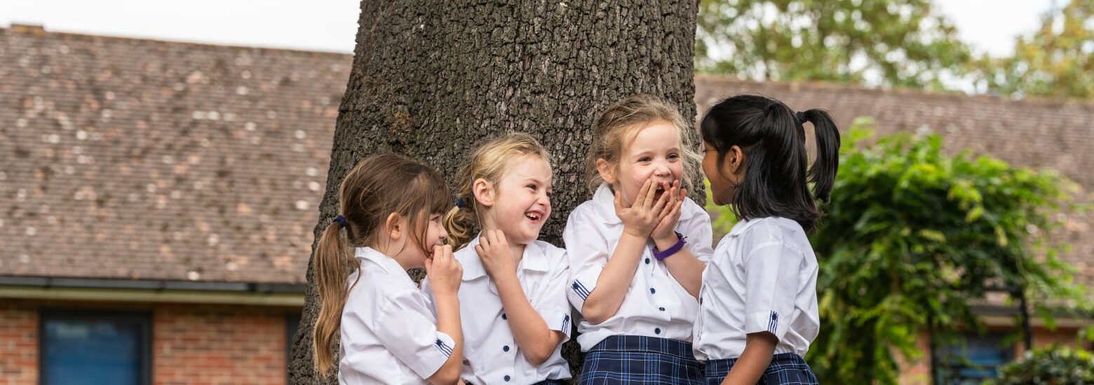 4 primary lower school girls in uniform standing by a tree talking and laughing about something one of the girls said at St Mary's school Open day
